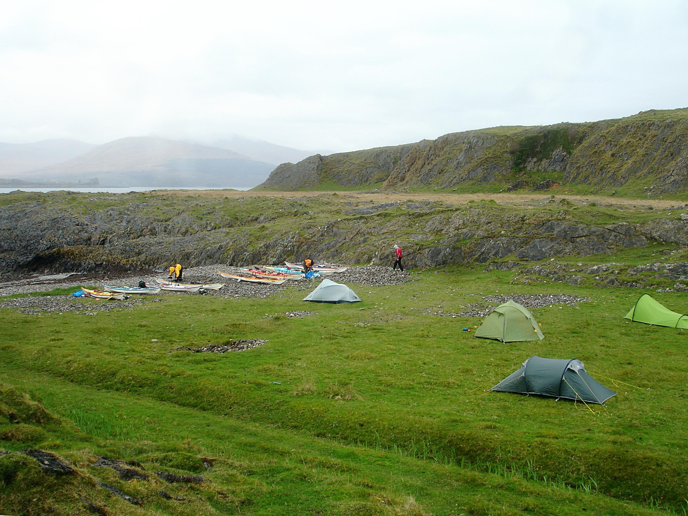 Multiple sea kayaks pulled up on a beach shoreline with tents set up at a multi-day paddling expedition camp