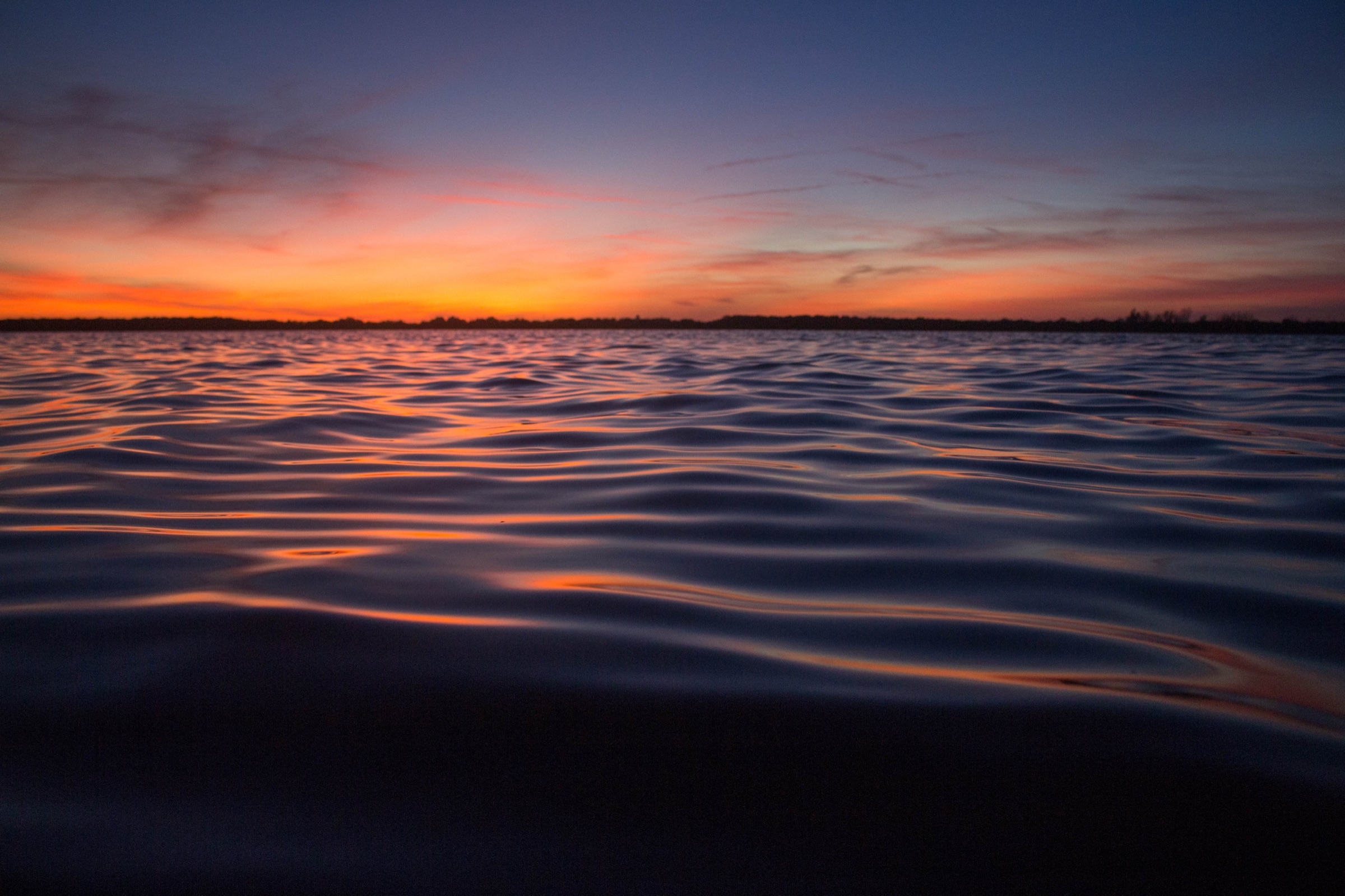 Vista ao nível da água da baía em Fort De Soto County Park, Flórida, sob um céu de pôr do sol laranja e roxo com a silhueta escura da vegetação no horizonte