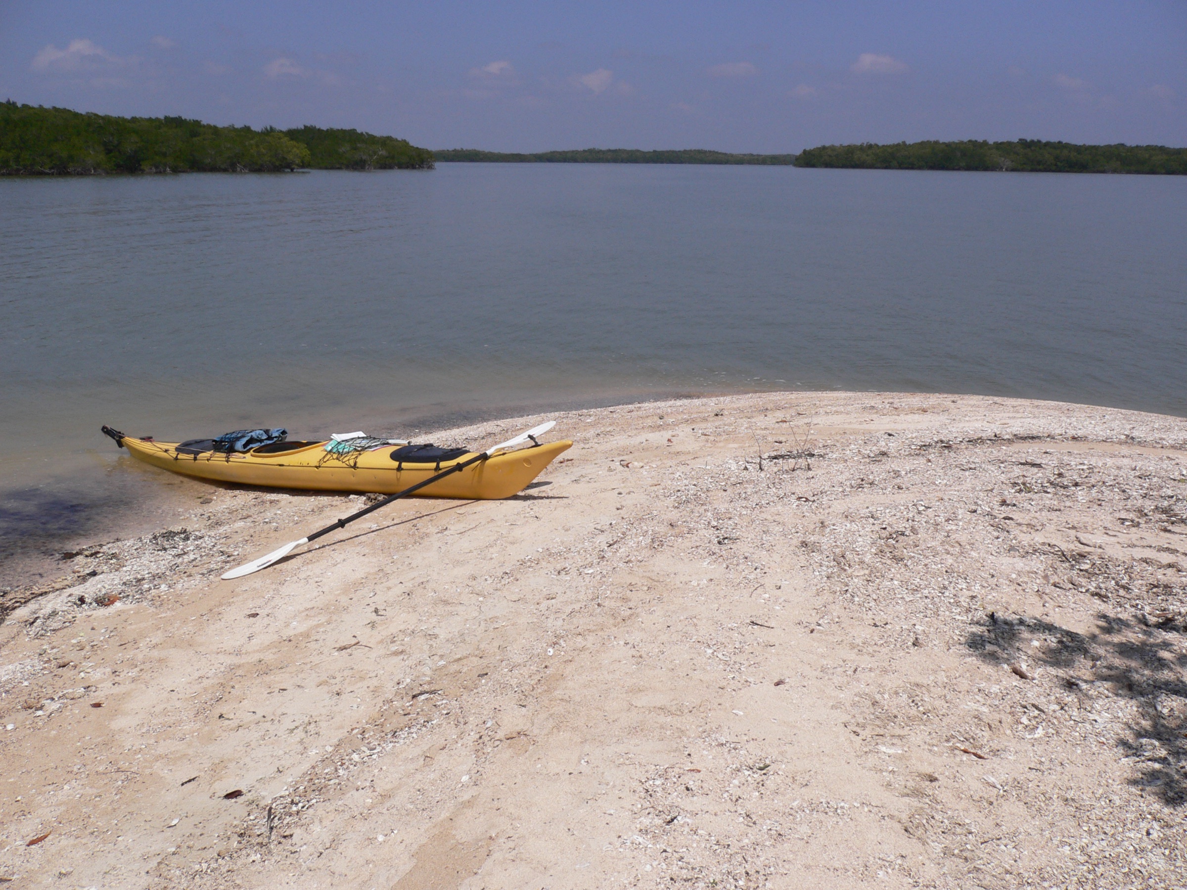 Un kayak de mar amarillo cargado con equipo descansa en una playa de arena de conchas en las Ten Thousand Islands de Florida, con aguas tranquilas e islas de manglar extendiéndose en la distancia