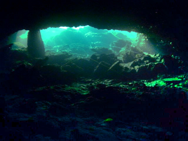 Underwater view from inside a Florida limestone spring cave, looking toward the glowing teal-lit cave entrance with rocky floor and walls at Peacock Springs