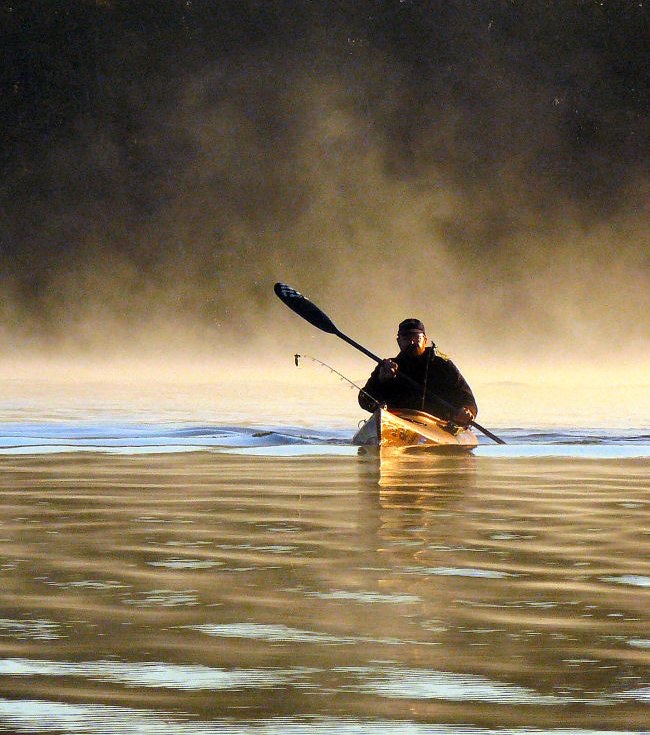 Angler fishing from a kayak on a misty lake at golden hour, paddle raised and fishing rod in hand