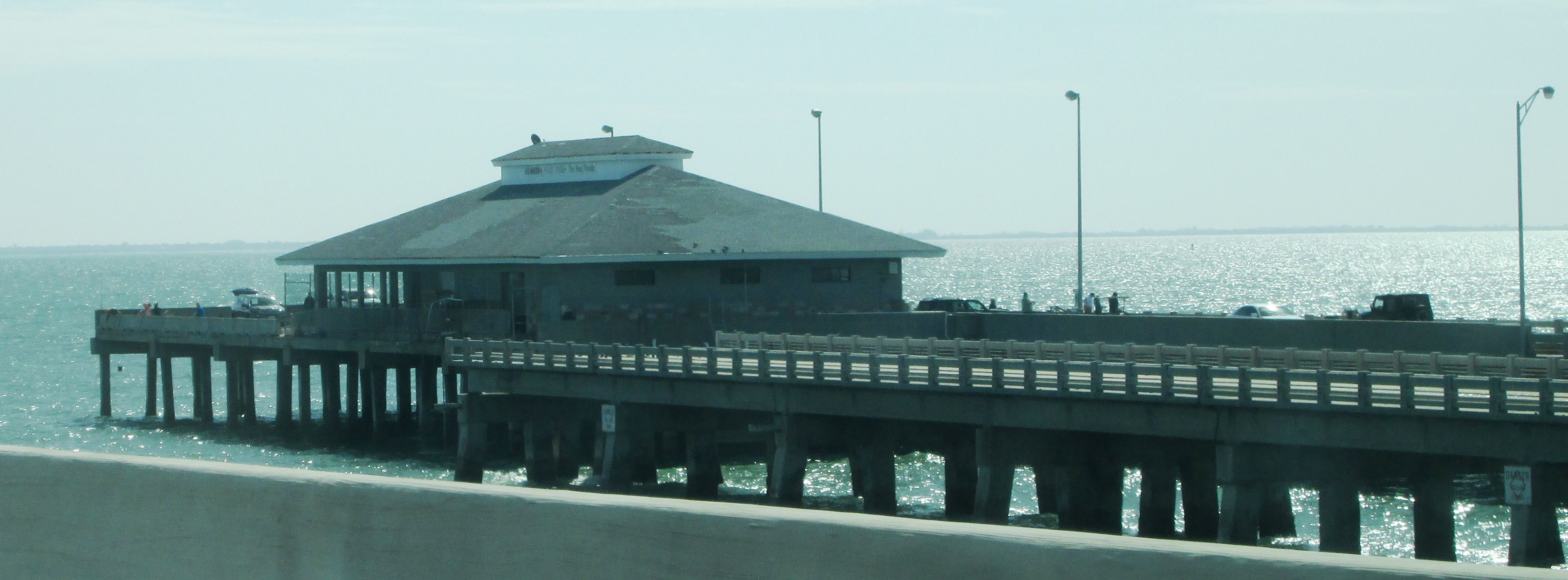 Aerial view of the long, low concrete fishing pier of Skyway Fishing Pier State Park stretching across Tampa Bay with cars parked along the rail and the cable-stayed Sunshine Skyway Bridge in the background