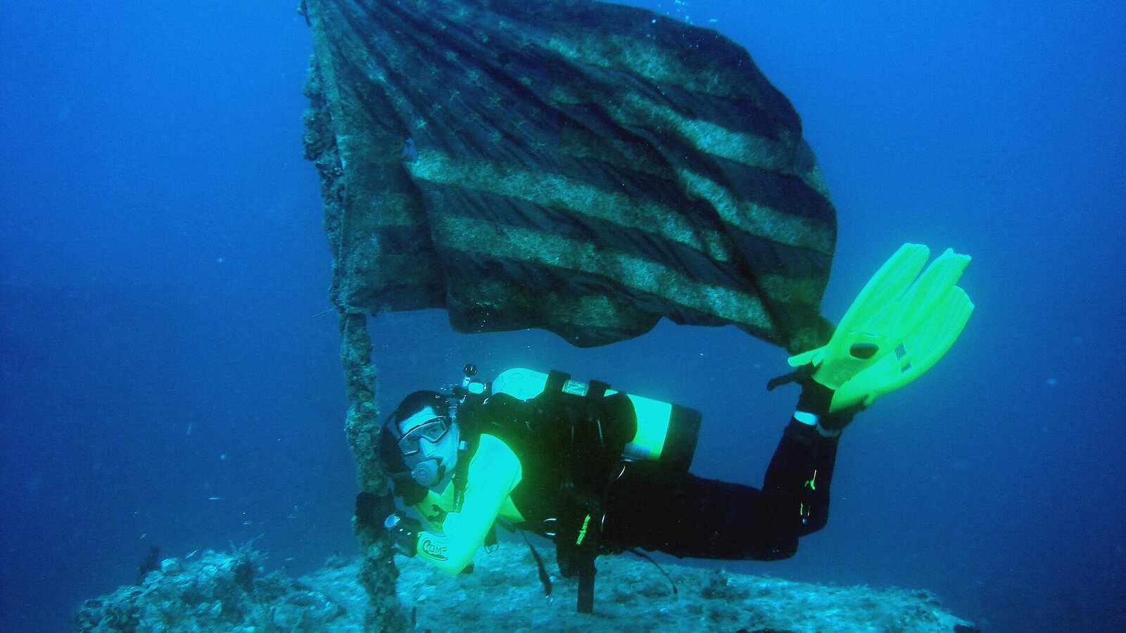 Diver descending toward the deck of a massive sunken naval ship covered in soft coral growth with shafts of sunlight from above